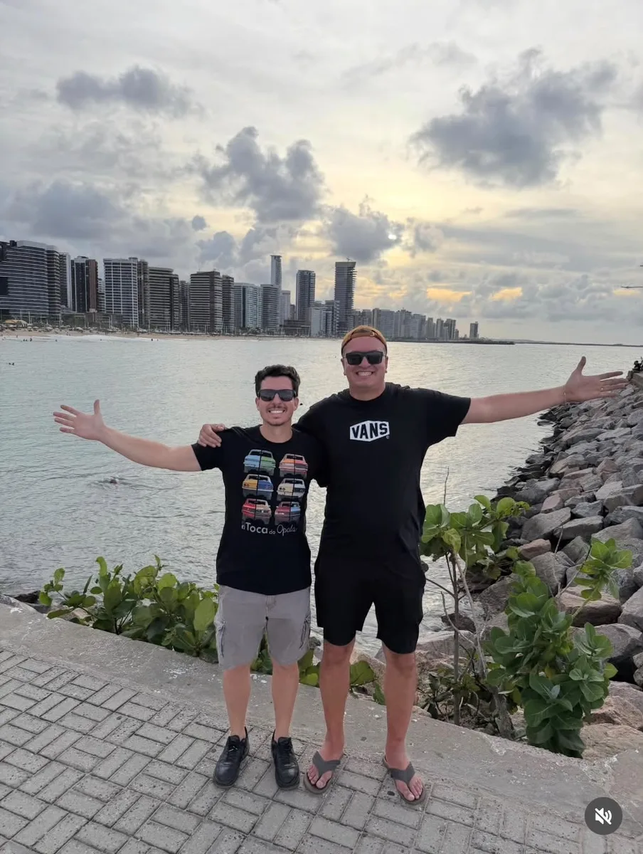 Os dois fundadores da Toca do Opala de braços abertos à beira-mar, com a cidade ao fundo